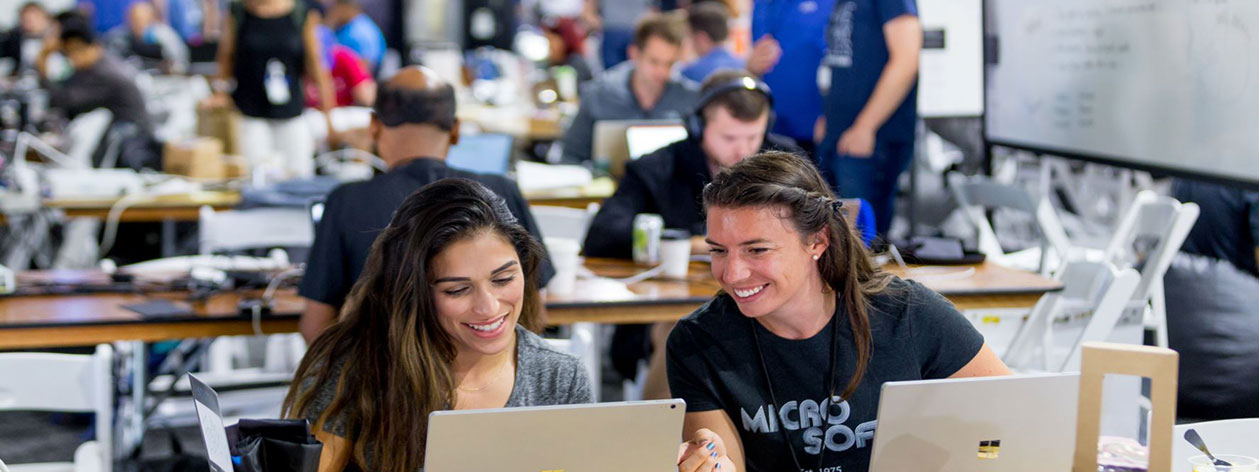 Two women using laptops confer while working together in a busy computer lab environment.