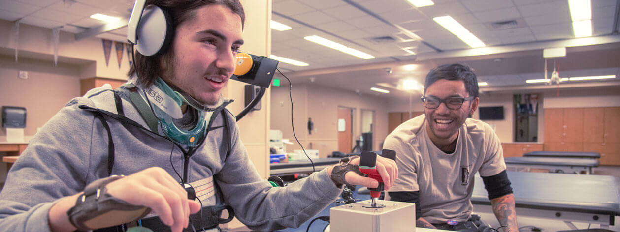 A man wearing arm and neck braces smiles while using enhanced controls to play a computer game