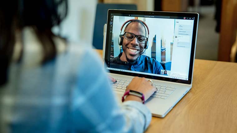 Woman at a desk using a Surface laptop to make a Microsoft Teams video call with one man smiling and wearing a headset.